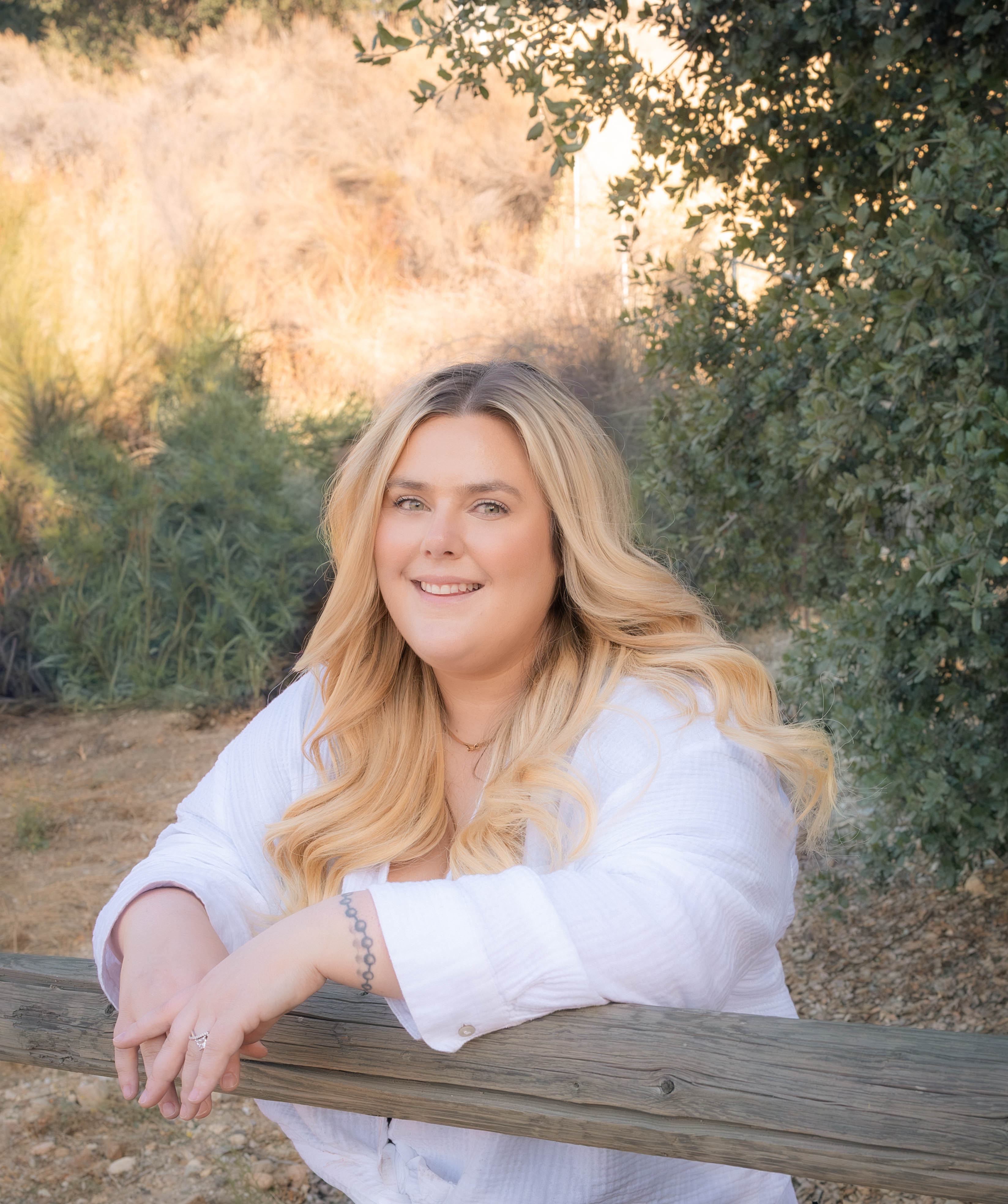 Ashley Kister leaning on a wooden fence, smiling, with warm golden-hour light in the background