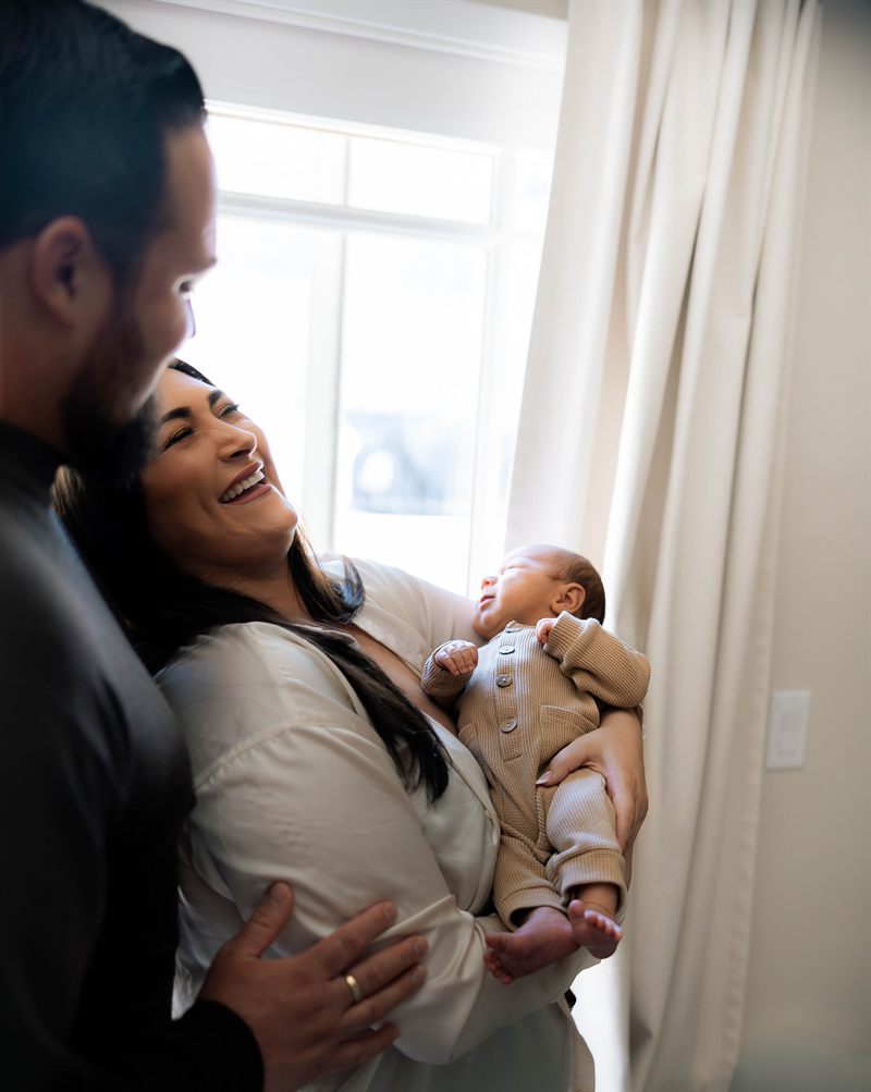 New parents laughing together while holding their newborn by a sunlit window